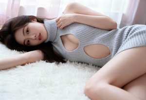 カットアウトのあるグレーのニットドレスを着て、リラックスしたポーズでふわふわの白い敷...。