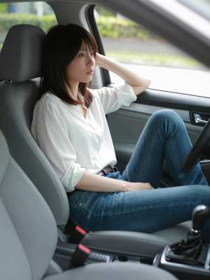 白いブラウスとブルー ジーンズを着て、現代の車の運転席に座っている若いアジア人女性を...。