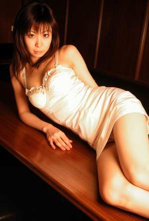 白いサテンのナイトガウンを着た若い女性が木の表面に座り、中立的な表情でカメラを直接見...。