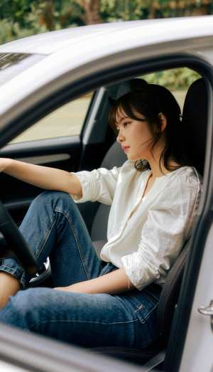 白いブラウスとブルー ジーンズを着て車の運転席に座っている若い女性を横から撮影した写...。