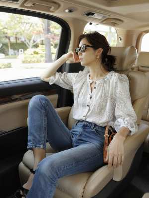 高級車の運転席に座り、ペイズリー柄の白いブラウスとブルー ジーンズを着て、黒いサンダ...。