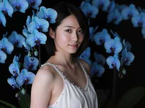 青い蘭の花の背景の前に立つ、白い肌と黒い髪の若い女性の写実的なポートレート。