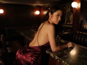 薄暗いバーのバーカウンターにもたれかかり、絹のようなワインレッドのドレスを着た若い女...。