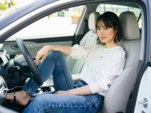 白いブラウスとブルー ジーンズを着て、現代の車の運転席に座り、リラックスした表情でカ...。