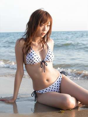 海を背景にビーチに座っている水玉模様のビキニを着た若い女性を横から撮った写実的な写真。