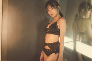 薄暗い部屋で鏡の前に立つ黒いランジェリーを着た若い女性を横から撮影した写実的な写真。