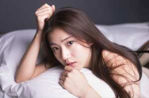 白いシーツが敷かれたベッドに横たわり、思慮深い表情で視聴者をまっすぐに見つめている若...。