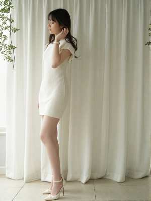 薄手の白いカーテンの前に立つ、白いニットドレスを着た妊婦の横からの写実的な撮影。