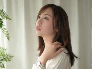 長い茶色の髪を持ち、白いセーターと時計を着て、白いカーテンと緑のシダ植物を背景にした...。