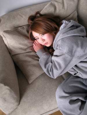 グレーのパーカーを着た若い女性がベージュのソファに座り、思慮深い表情でカメラを直接見...。