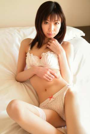 白いランジェリーのアンサンブルを着てベッドに座り、瞑想的な表情でポーズをとる若い女性...。