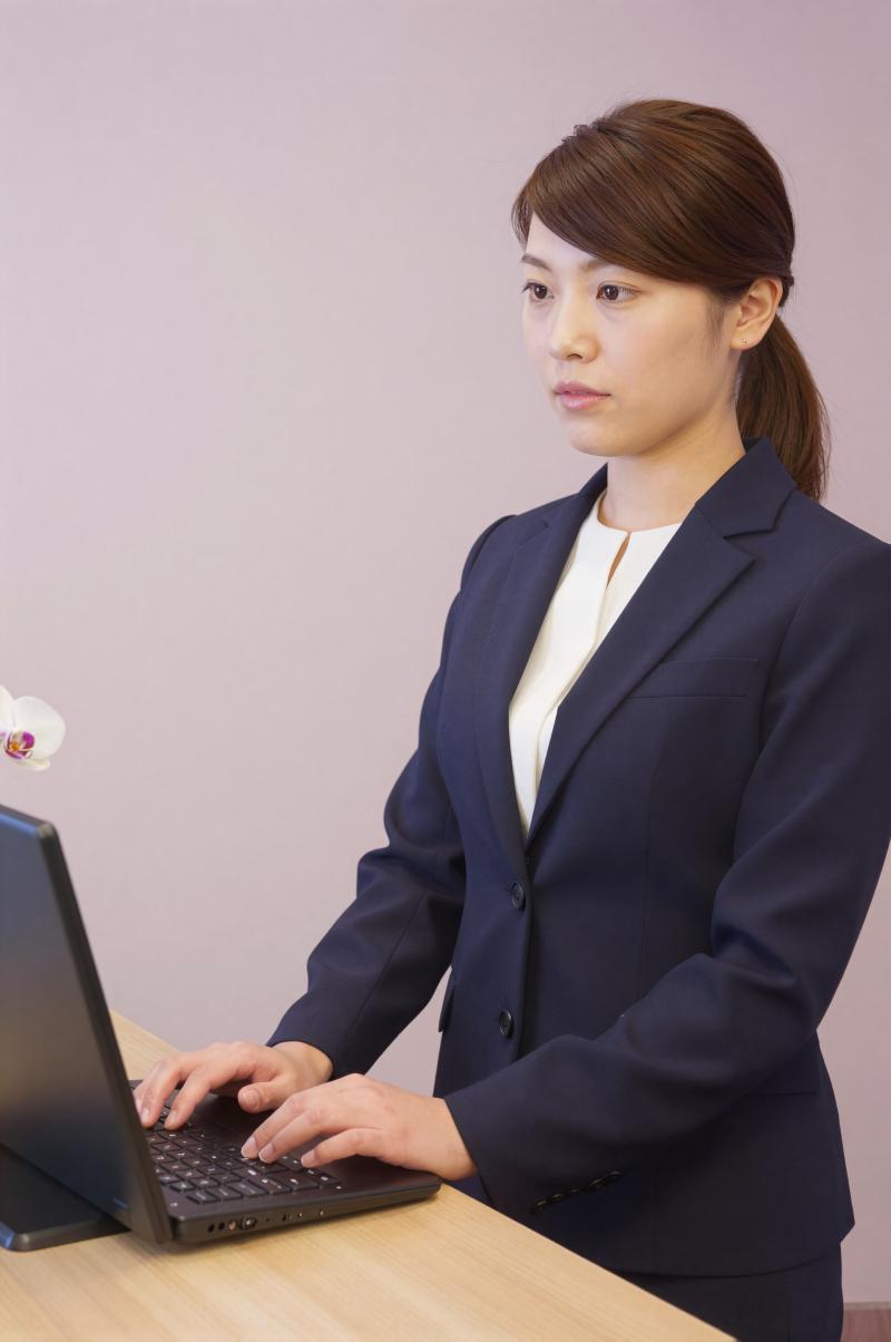 ホテル受付でPC作業と来客対応する女性