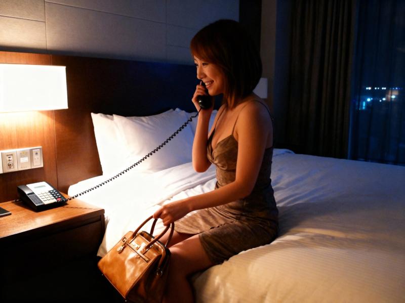 ホテルの部屋のベッドに座り、茶色のハンドバッグを持ちながら電話で話している女性。