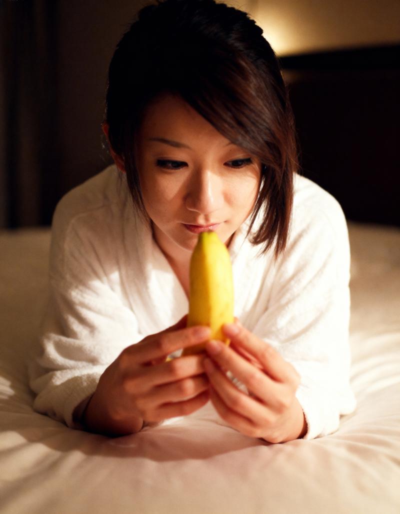 ベッドに座って顔の前に黄色いバナナを持ち、瞑想的な表情でそれをじっと見つめている若い...。