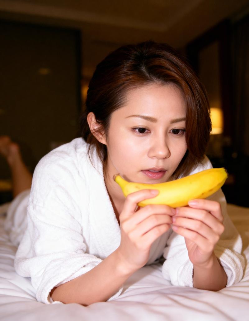 白いバスローブを着た若い女性がベッドに横たわり、顔の前にバナナを持っている様子を、ク...。