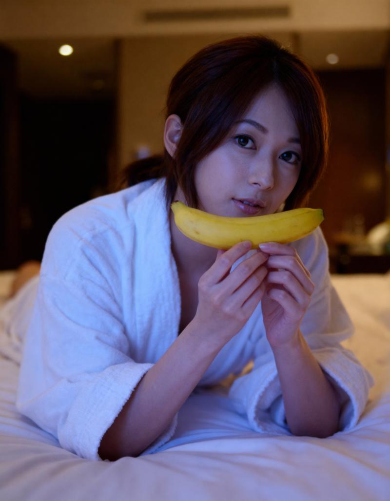 白いバスローブを着た若い女性がベッドに横たわり、顔の前にバナナを持ち、無表情でカメラ...。