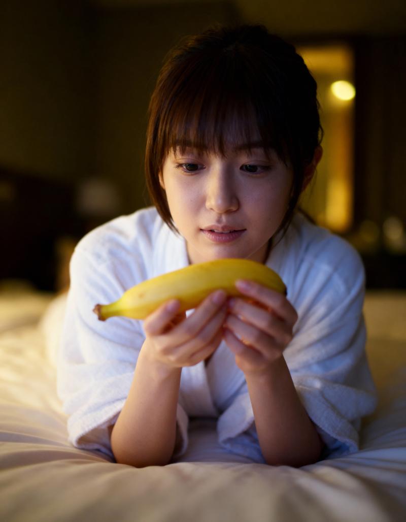 薄暗い寝室のベッドに横たわり、顔の前にバナナを持った若い女性をクローズアップ カメラ...。