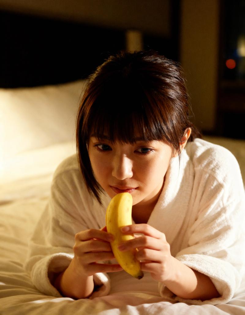 ベッドに座ってバナナを持ち、無表情でカメラを見つめる若い女性を正面から写実的に撮影したもの。