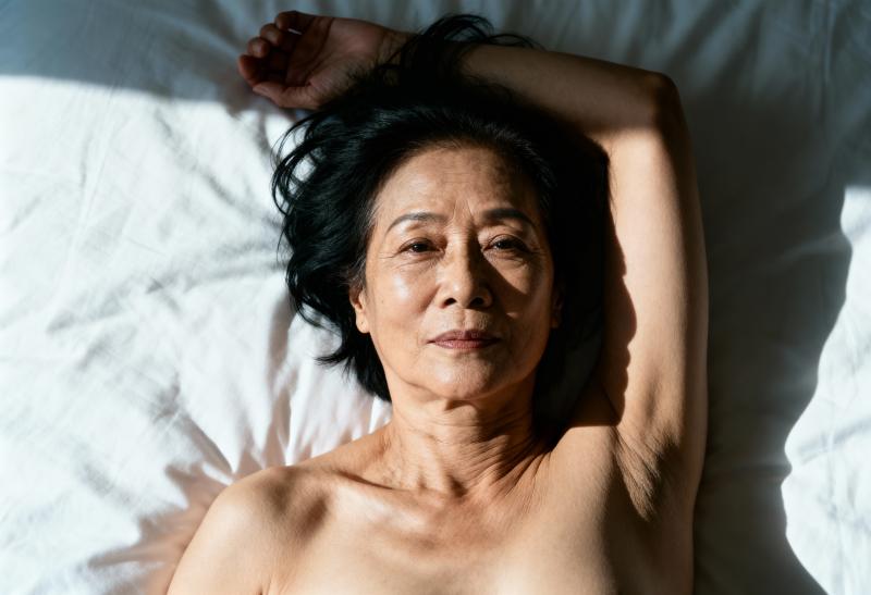 白いシーツが敷かれたベッドに横たわり、上からカメラを直接見つめている年配のアジア人女...。