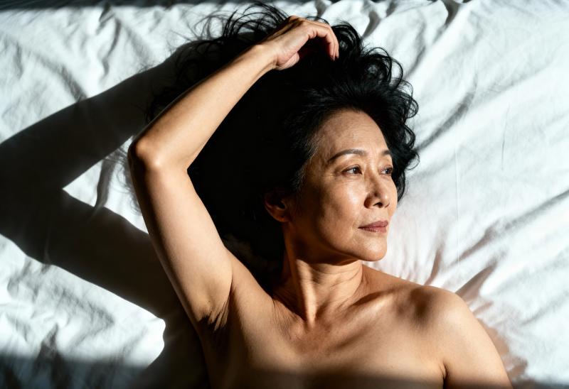 白いベッドシーツの上に横たわり、右腕を横に上げ、左腕を頭の上に置いている中年のアジア...。