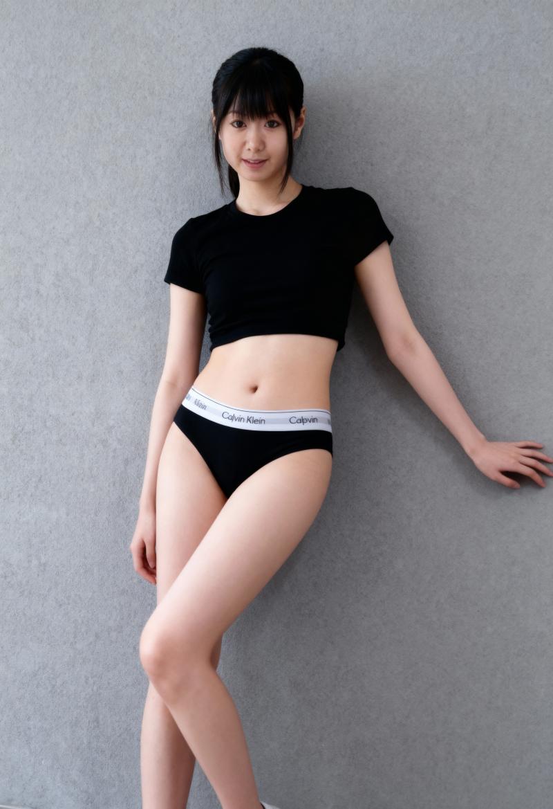ミニマルな環境の中で、最小限の服を着て灰色の壁に向かってポーズをとる若い女性を横から...。