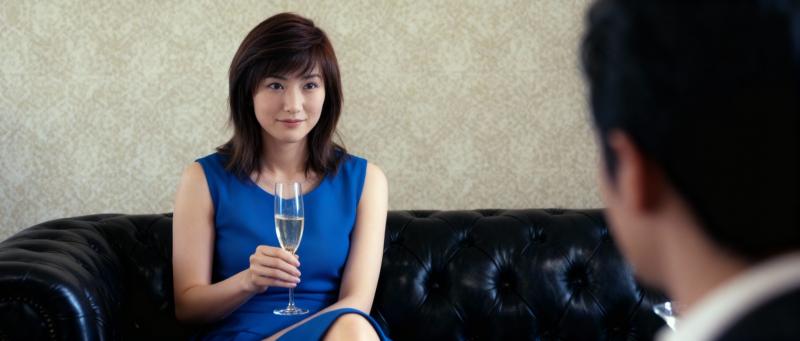 青いドレスを着た女性がシャンパンのグラスを持って黒い革張りのソファに座り、背景の男性...。