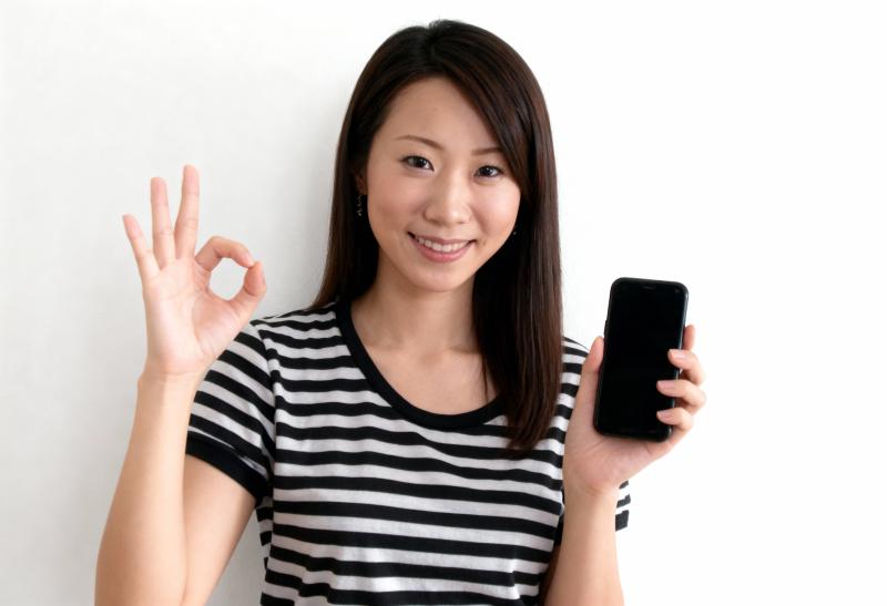 無地の白い背景の前に立ち、右手に黒いスマートフォンを持ち、左手で OK サインを出し...。