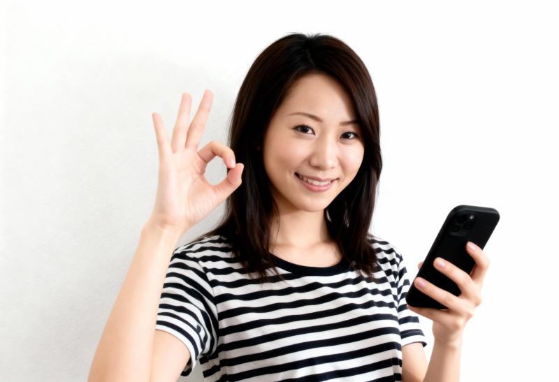 無地の白い背景の前に立ち、右手に黒いスマートフォンを持ち、左手で「OK」のジェスチャ...。
