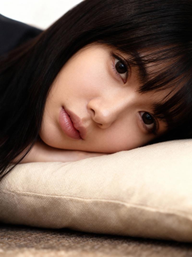 ベージュの枕に横たわり、穏やかな表情でカメラをまっすぐ見つめている若い女性の接写。