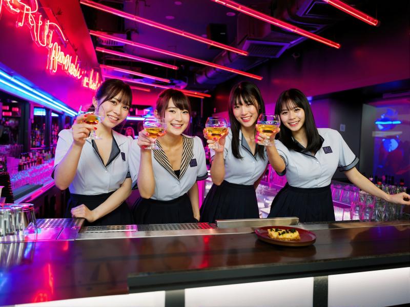 ネオンが灯る薄暗いバーで、バーの後ろに立っている 4 人の若い女性がそれぞれ異なる色...。