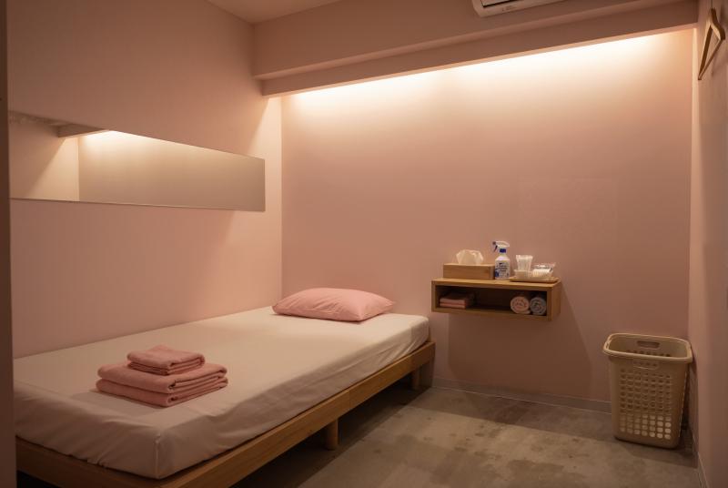 木のフレームと淡いピンクの壁が部屋の中央にシングルベッドを置いた、ミニマルなデザイン...。