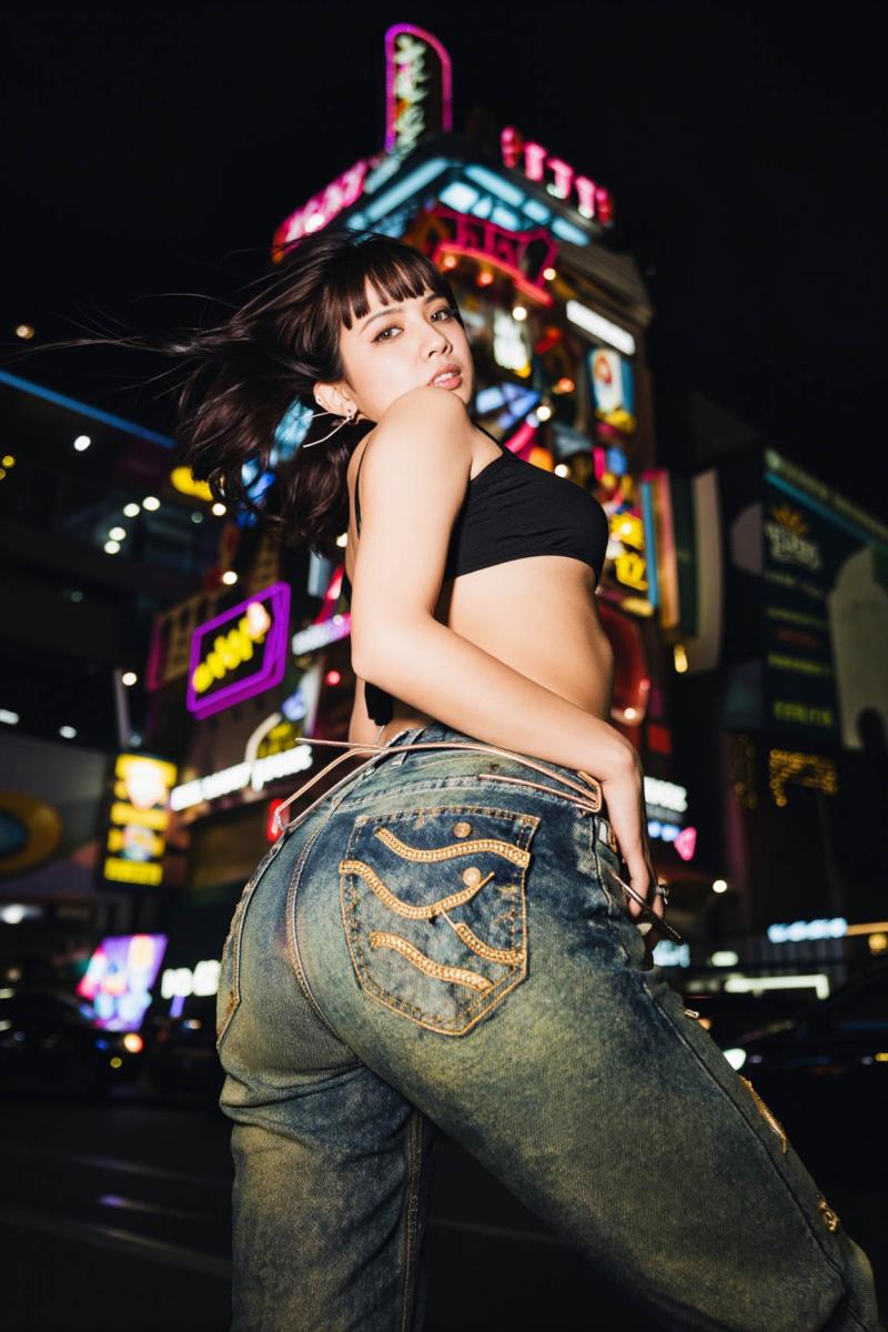 夜のネオン輝く街並みの中で黒いクロップトップとジーンズを着てポーズをとる女性を後ろか...。