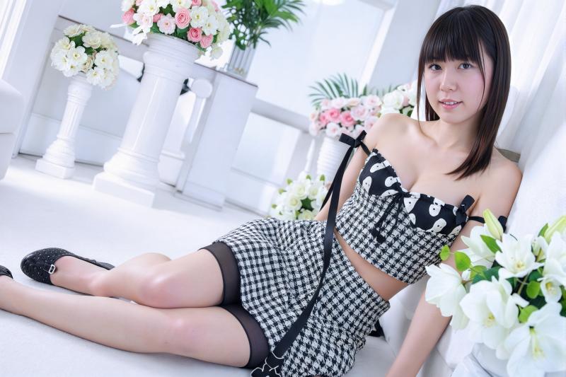 白い部屋で花に囲まれた床に座る若い女性を正面から写実的に撮影した写真。